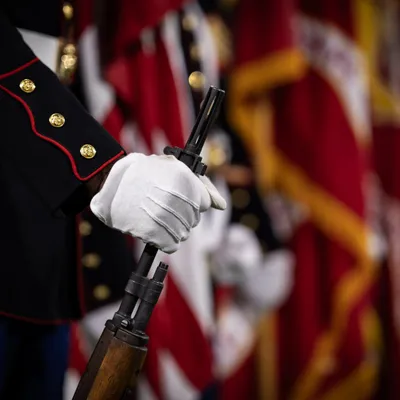 A white-gloved hand grips a rifle in front of the unit colors